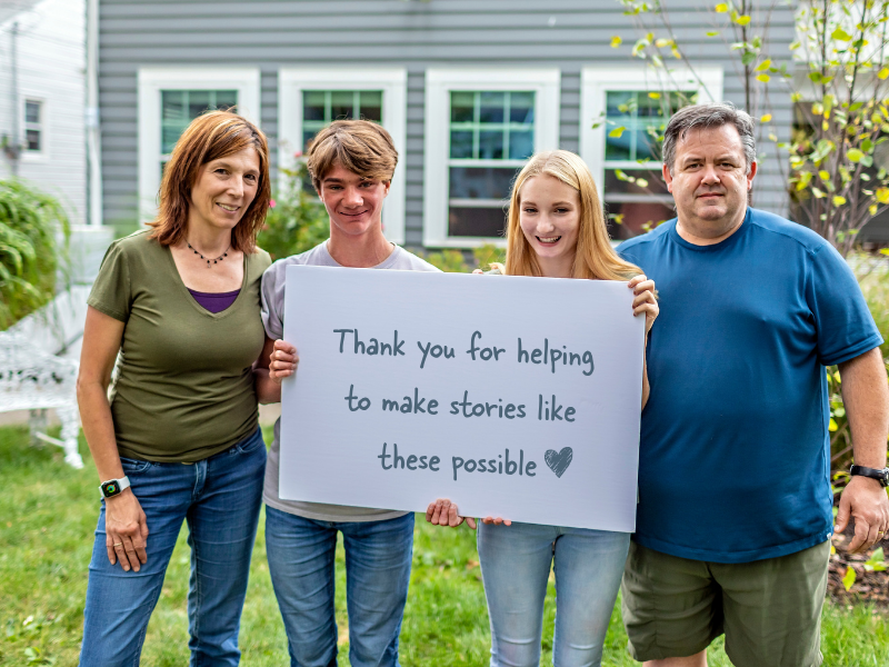 Four people stand outdoors smiling, with two in the middle holding a sign that says, Thank you for helping to make stories like these possible with a heart symbol at the end. A house and greenery are in the background.