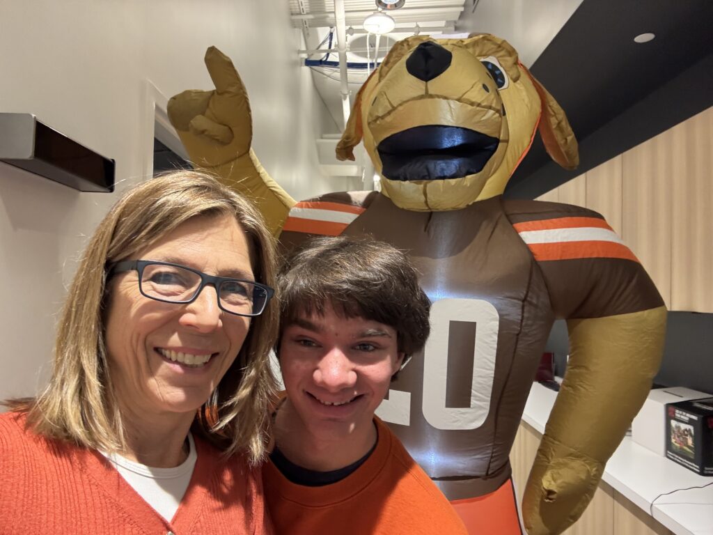 A woman and a teenage boy smile for a selfie indoors, standing in front of a large inflatable dog mascot wearing a football jersey with the number 20.
