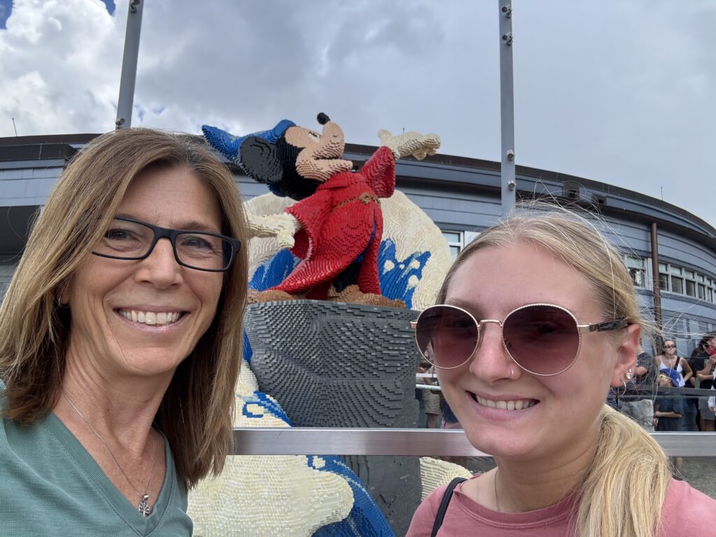 Two women smiling in front of a large LEGO sculpture of Mickey Mouse dressed as a wizard. The background shows a crowd and a modern building, with a cloudy sky above.