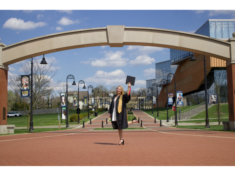 A graduate in a black cap and gown stands under a large stone arch on a university campus, holding up her diploma. Modern buildings and banners line the walkway. The sky is blue with scattered clouds.