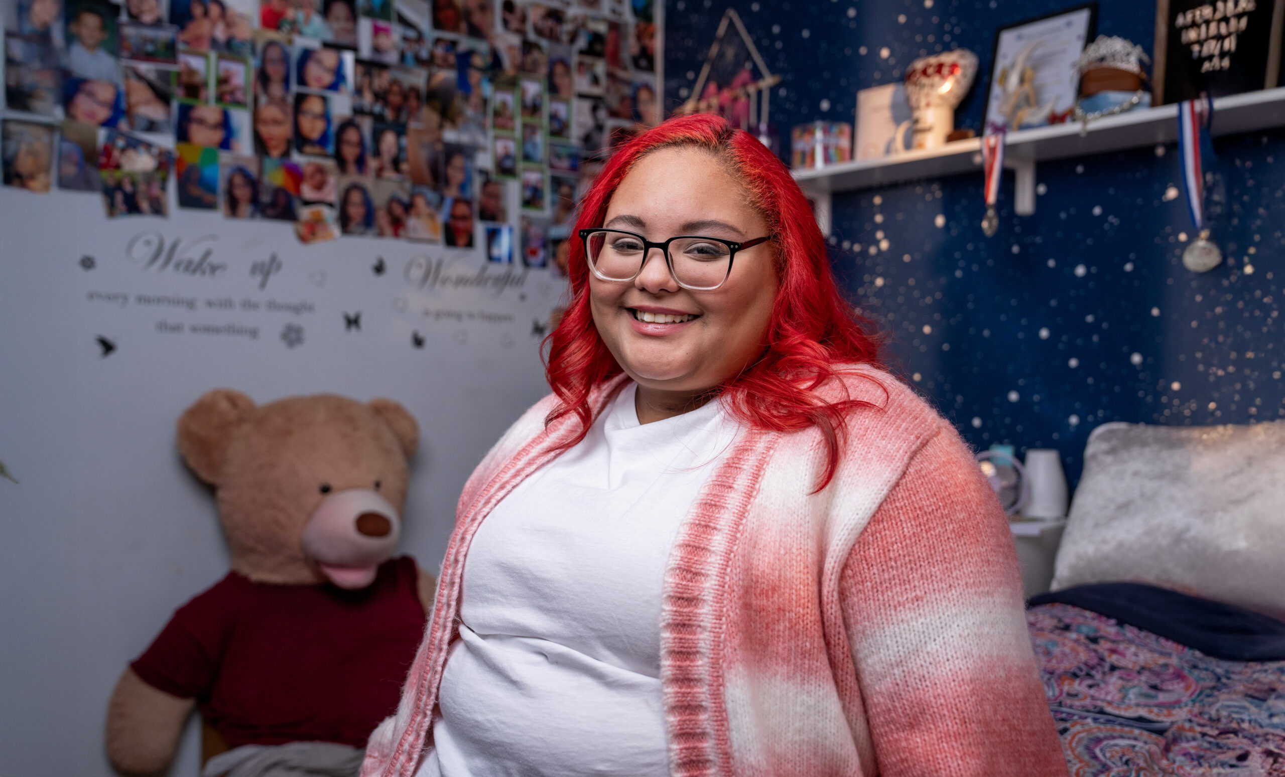 A teenage girl with red hair and glasses sits in a cozy bedroom with a large teddy bear, a photo collage on the wall, and starry wall decor. They wear a white shirt and pink cardigan.