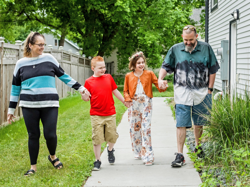 A family of four walking hand-in-hand along a concrete sidewalk beside a house.
