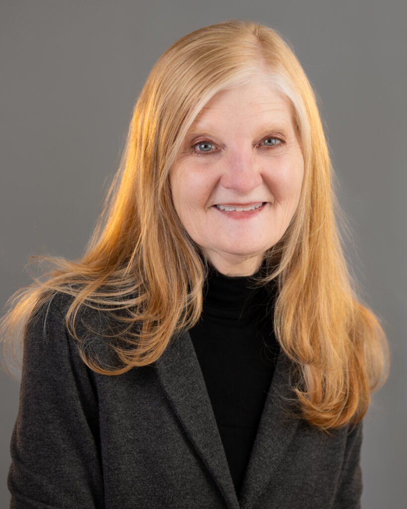 A smiling older woman with long blonde hair wearing a dark blazer and black top poses in front of a plain gray background.