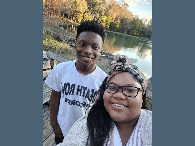 A smiling woman and a boy take a selfie outdoors on a wooden deck by a calm lake, with colorful autumn trees in the background under a blue sky.