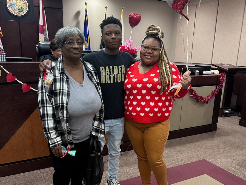 Three people stand and smile in a decorated room with heart-shaped balloons and garlands. One woman wears a plaid jacket, the young man wears a Raider Nation shirt, and another woman wears a red sweater with white hearts.