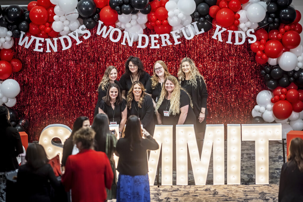 A group of women pose and smile in front of a red and black balloon arch and a sparkling red backdrop with Wendys Wonderful Kids SUMMIT in large lit-up letters. People in the foreground take photos.