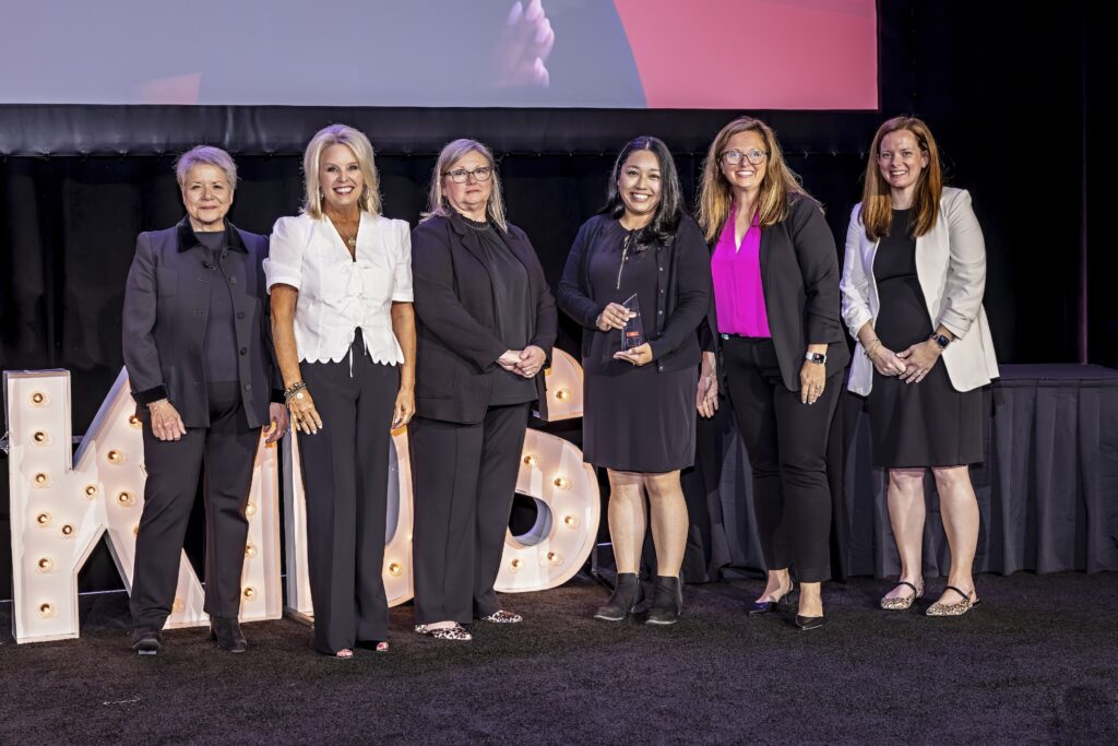 Six women stand together on a stage, five in black outfits and one in a white jacket. One woman in the middle holds an award. Large lit-up letters are visible behind them. All are smiling and facing the camera.