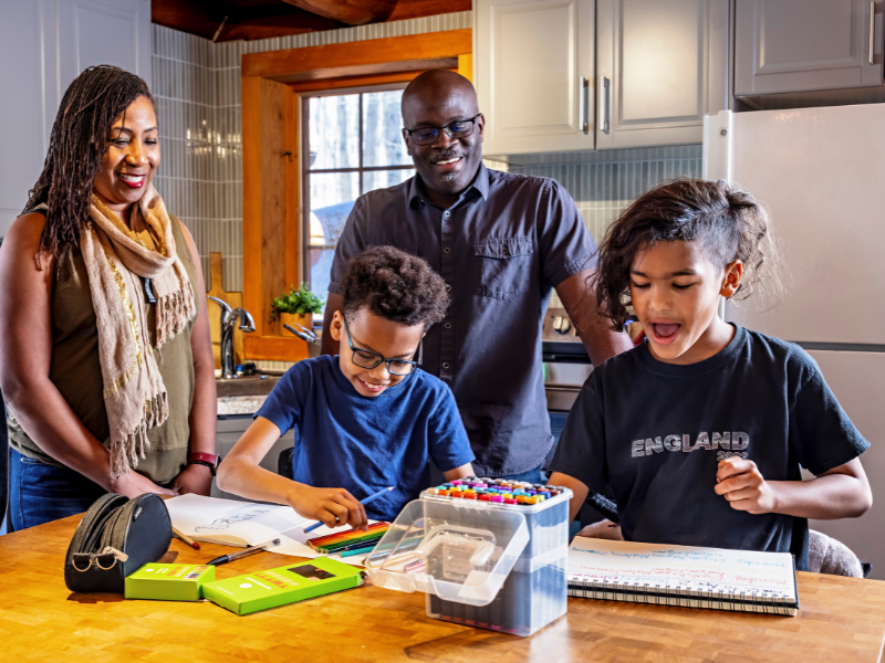 A smiling family of four stands in a kitchen. Two children draw with colored pencils at a wooden table while two adults watch happily. Art supplies and sketchbooks are scattered on the table.