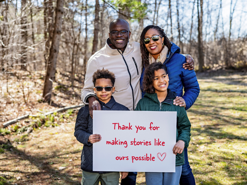 A smiling family of four stands outdoors in a wooded area, holding a sign that reads, Thank you for making stories like ours possible with a heart drawn next to the text.