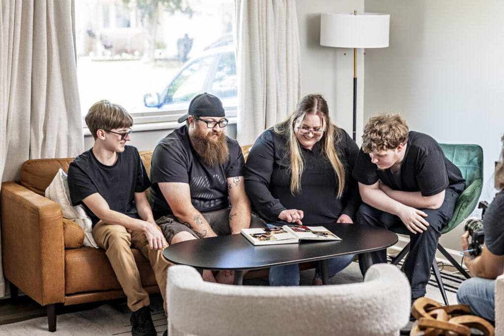 A group of four people sit around a coffee table in a living room, smiling and looking at an open photo album together. They appear relaxed and engaged in their shared activity.