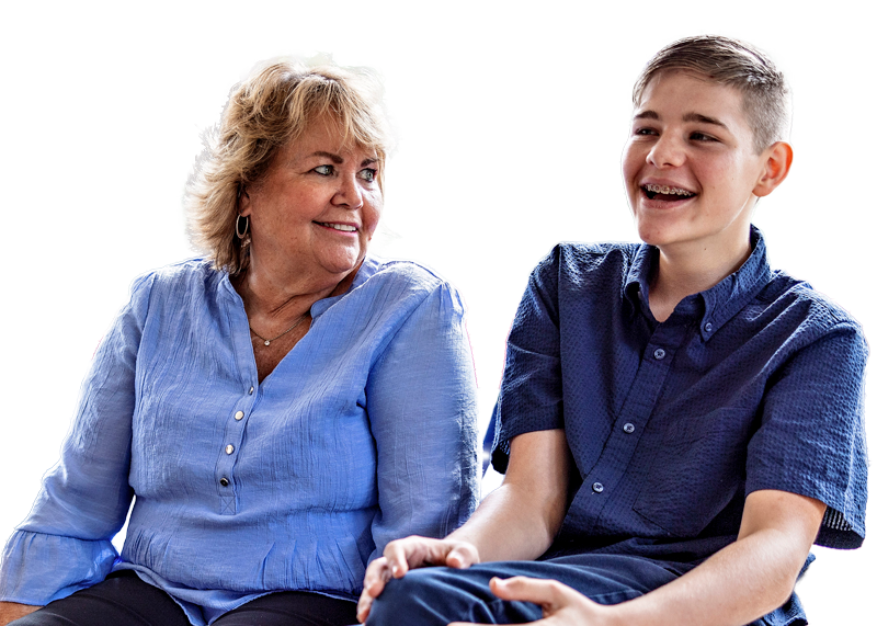 An older woman and a teenage boy sit next to each other, smiling and laughing. Both are wearing blue shirts and appear happy and engaged, with a white background behind them.