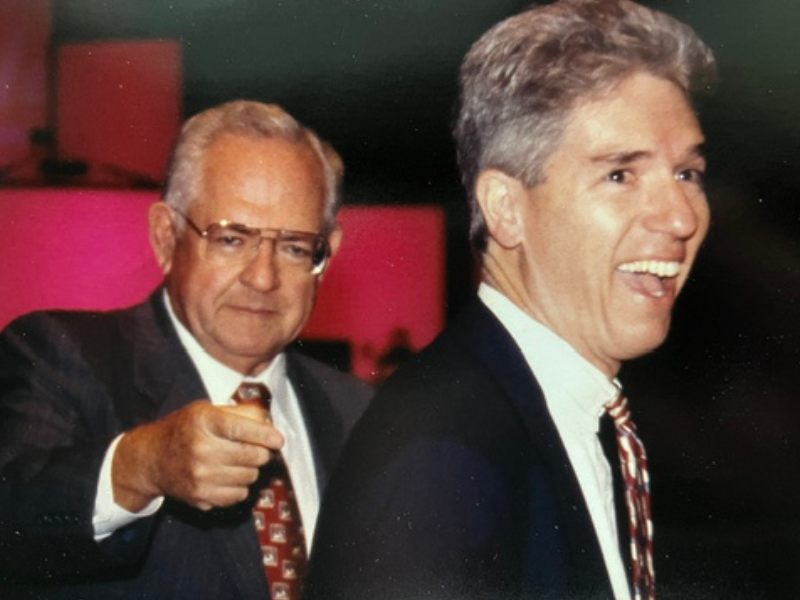 Two men in suits, one smiling broadly in the foreground, the other behind him looking serious and pointing forward.