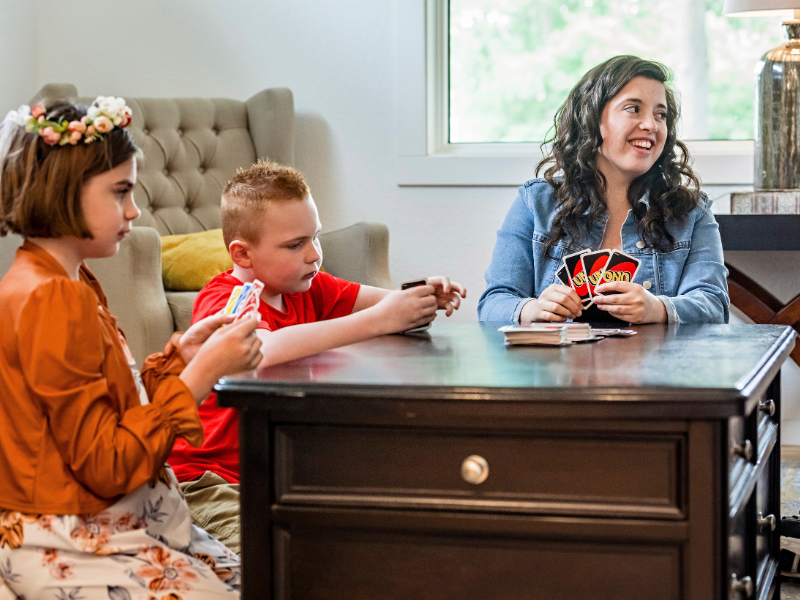 A woman and two children sit around a dark wooden table in a bright room, playing a game of Uno. The girl on the left wears a floral crown, a boy in a red shirt focuses on his hand, and the woman on the right smiles while holding her cards.