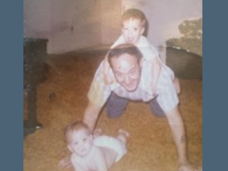 A man on all fours with a smiling child on his back and a baby in a diaper crawling on the carpet in front of him.