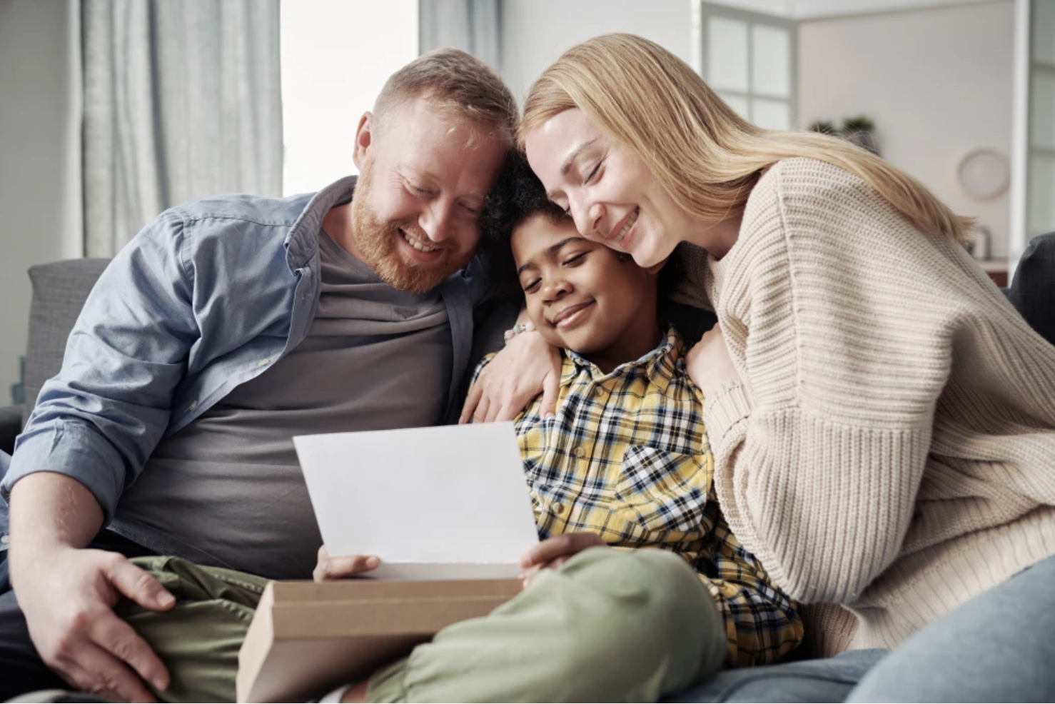 A smiling family of three sits close together on a couch, embracing as they look at a piece of paper or card. The child is in the middle, held lovingly by the two adults.