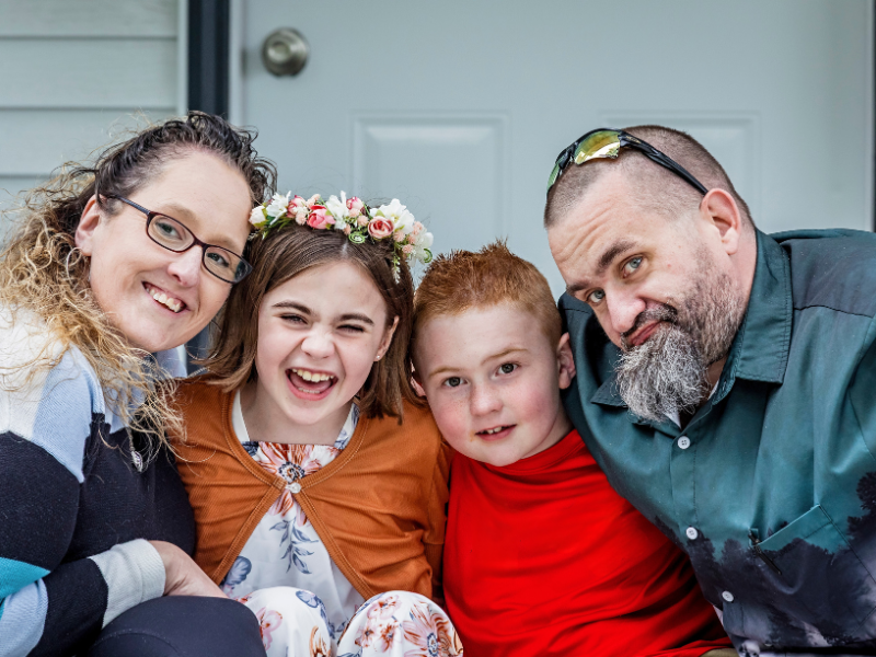 Four people—two adults and two children—sit closely together, smiling. One girl wears a floral crown and an orange wrap, while the boy has short red hair and wears a red shirt. They all look happy in front of a white door.