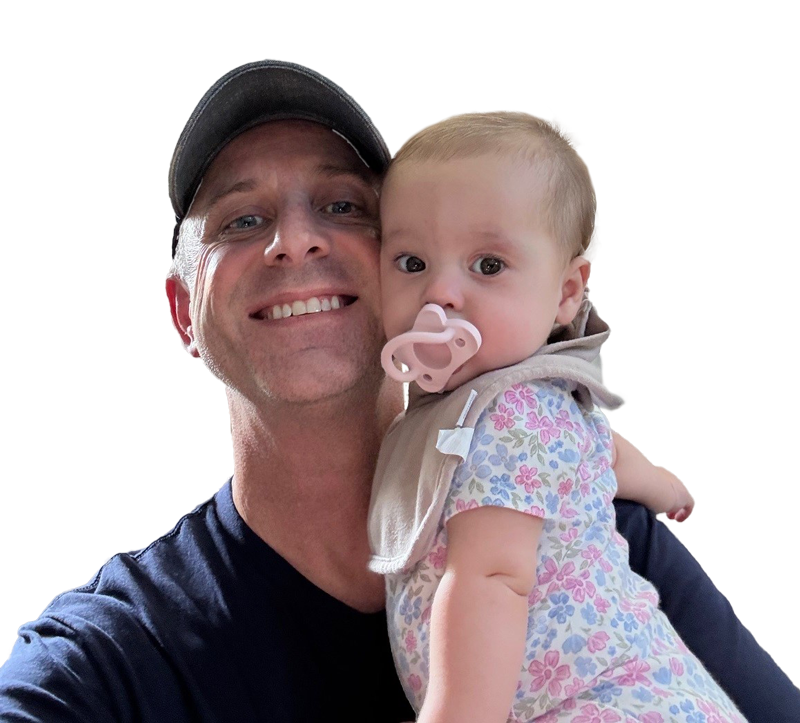 A smiling man in a dark cap holds a baby with a pink pacifier and floral outfit. The background is plain white.