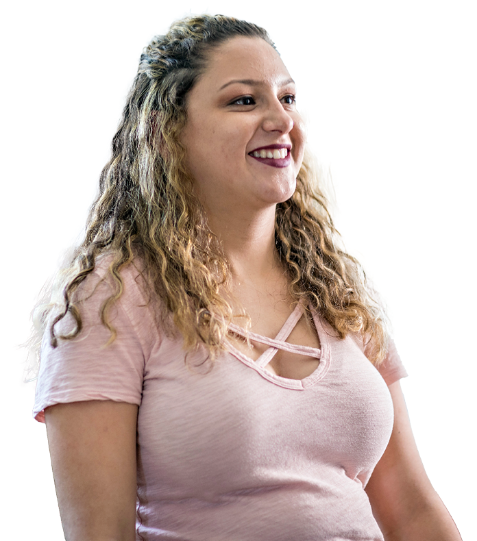 Older youth with curly, light brown hair wearing a pink short-sleeve top smiles while looking slightly to the side against a plain white background.