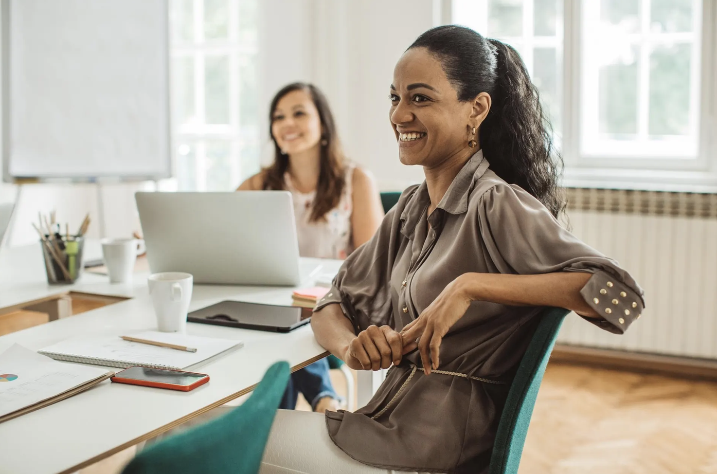 Two women in an office setting are smiling; one sits at a desk with a laptop, papers, and coffee mugs, while the other sits in the foreground, looking to the side. Bright windows provide natural light in the background.