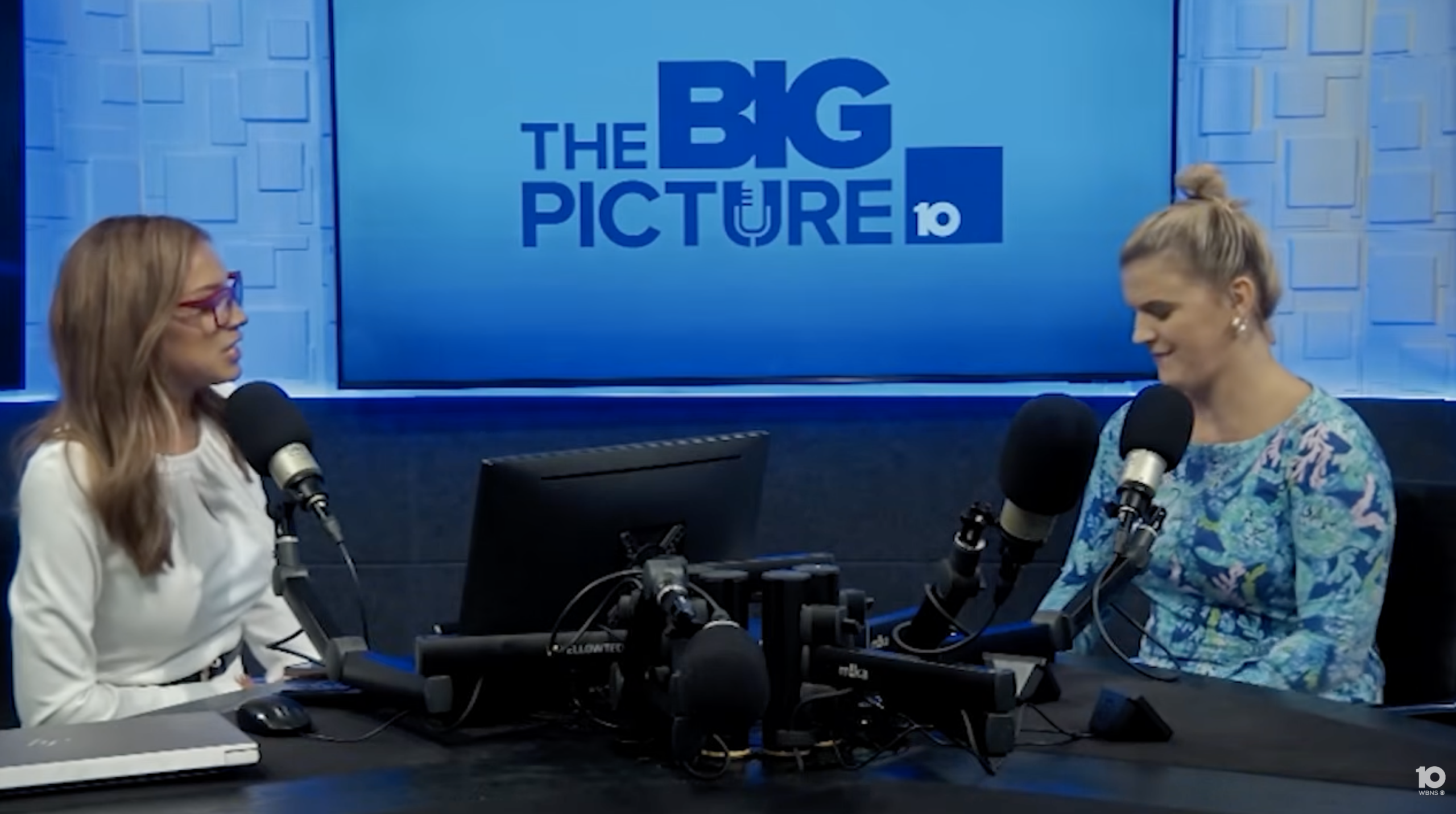 Two women sit in a radio studio with microphones, one in a white top and glasses, the other in a blue patterned dress, beneath a large sign reading The Big Picture 10..