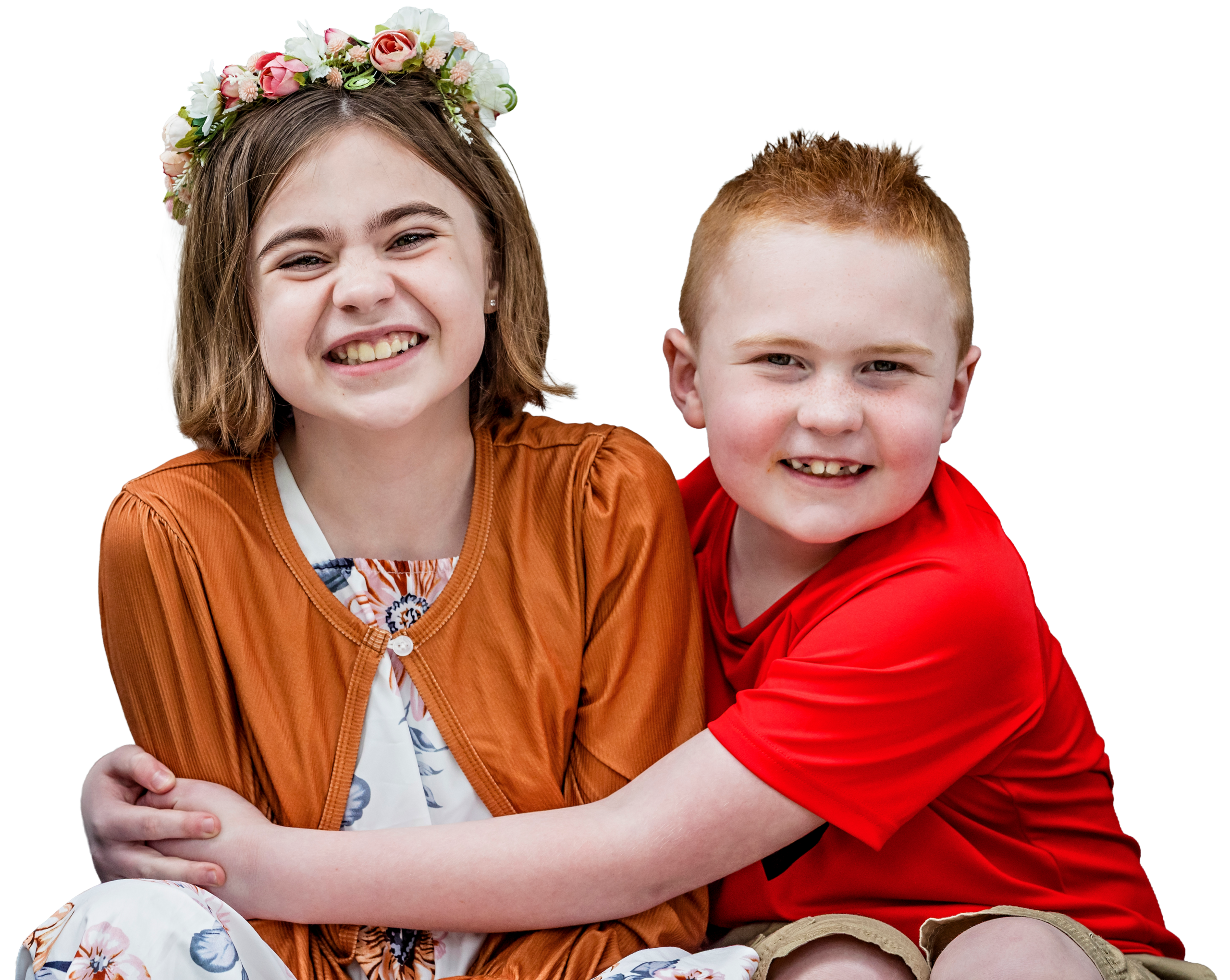 Two smiling children pose together. The girl on the left wears a orange cardigan, floral dress, and a flower crown, while the boy on the right in a red shirt hugs her from the side.