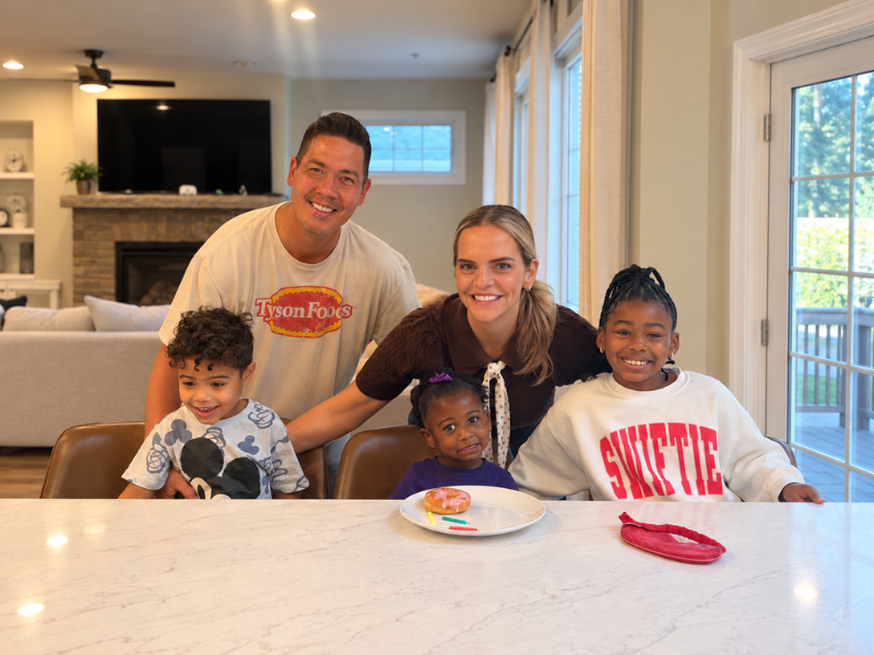 A smiling family of five poses at a kitchen counter. Three children sit in front, with two adults standing behind them. There is a plate with a donut and a crayon on the table. Bright windows are in the background.