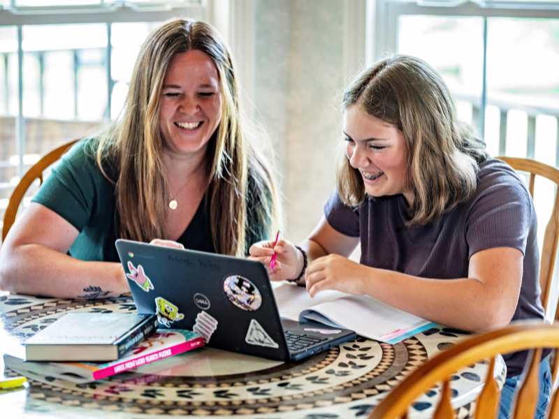 Two young women sit at a table smiling while working together on a laptop and taking notes, with books and pens spread out in front of them in a brightly lit room.
