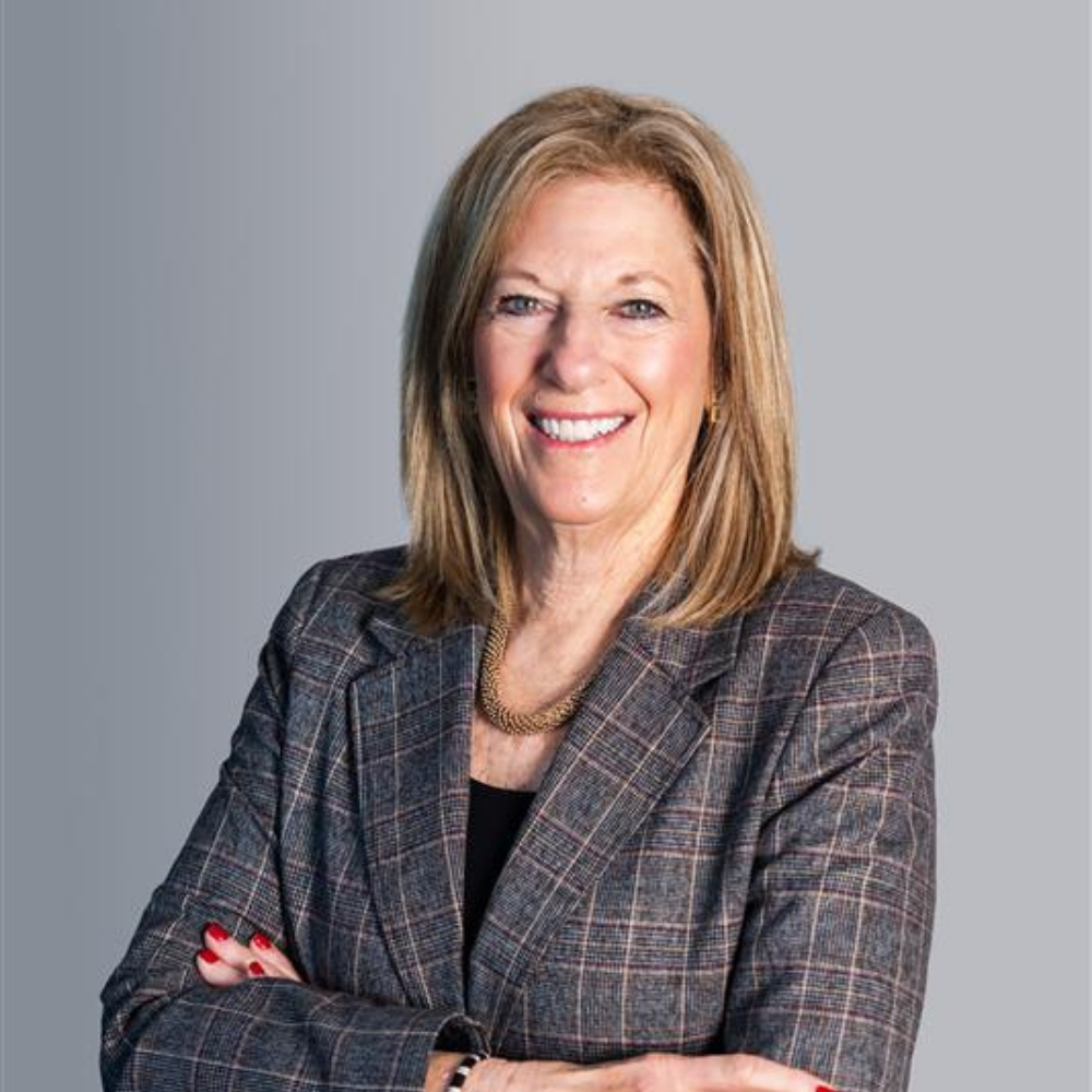 A smiling woman with shoulder-length light brown hair wears a plaid blazer over a black top, a necklace, and has her arms crossed. The background is a plain light gray gradient.
