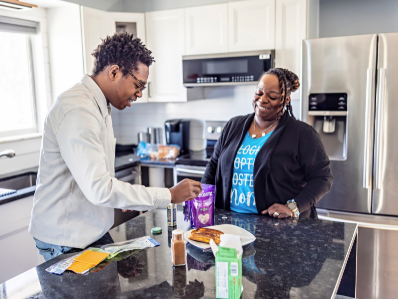 A mother and son smile in a modern kitchen as he prepares food at the counter. Various ingredients are spread out by a plate of toast. Sunlight streams through the window.