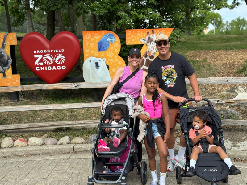A smiling family poses at Brookfield Zoo Chicago in front of a colorful animal sign, with two young children in strollers and an older child standing between two adults.