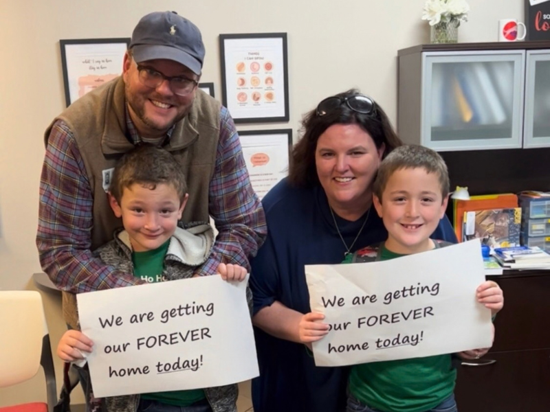 A smiling family of four poses indoors as two children hold signs that read, We are getting our FOREVER home today!.
